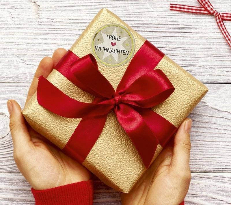 Hände halten ein in Gold eingewickeltes Geschenk mit einer roten Schleife und Schleifchen, mit der Aufschrift „FROHE WEIHNACHTEN