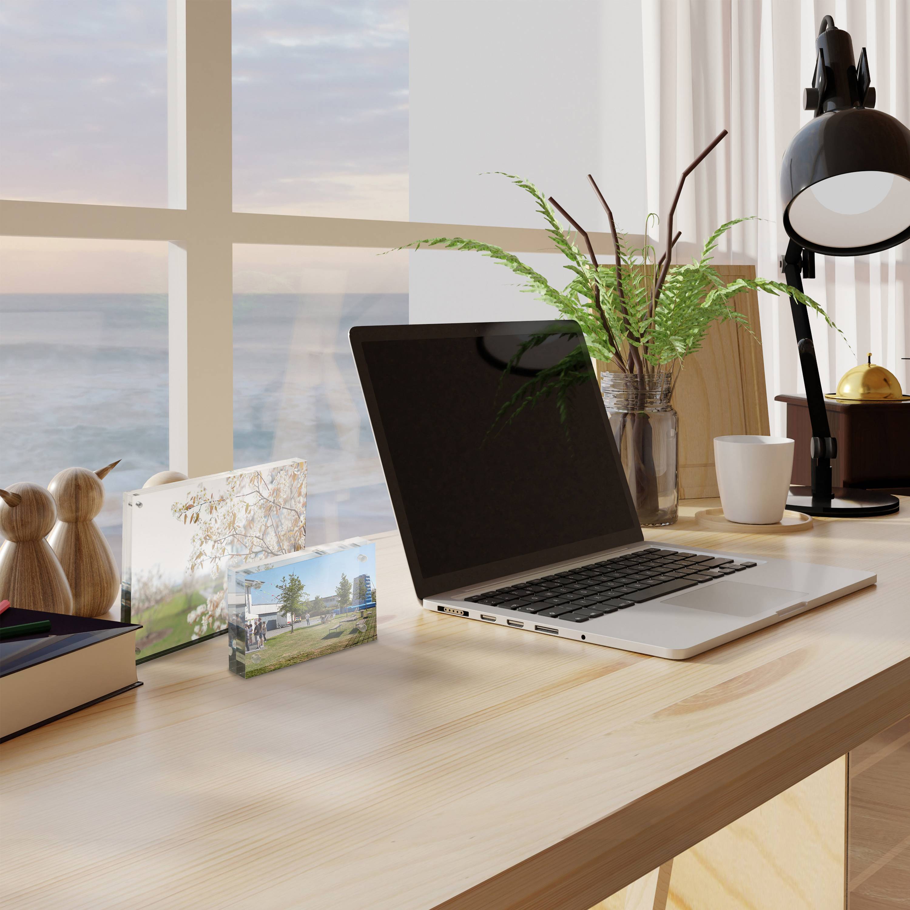 Laptop auf einem hellen Schreibtisch neben einem Fenster, mit einer Pflanze, Schreibtischlampe, Tasse, Büchern und dekorativen Fotokarten in einem gemütlichen Arbeitsbereich.