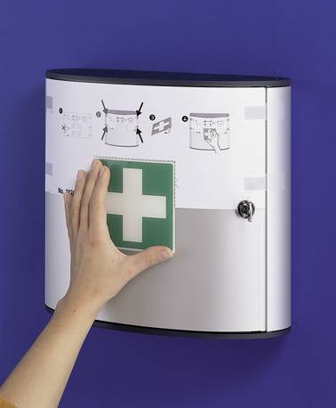 Hand platziert ein grünes Erste-Hilfe-Kreuzzeichen auf einem weiß wandmontierten medizinischen Schrank mit Anweisungssymbolen auf violettem Hintergrund.