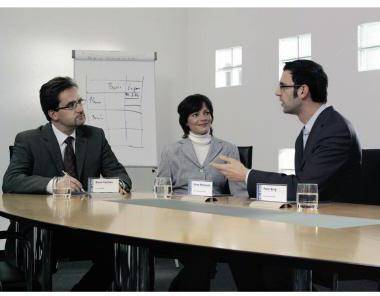 Drei Geschäftsleute in einem Konferenzzimmer, wobei ein Mann zu den anderen über einen Tisch hinweg spricht.