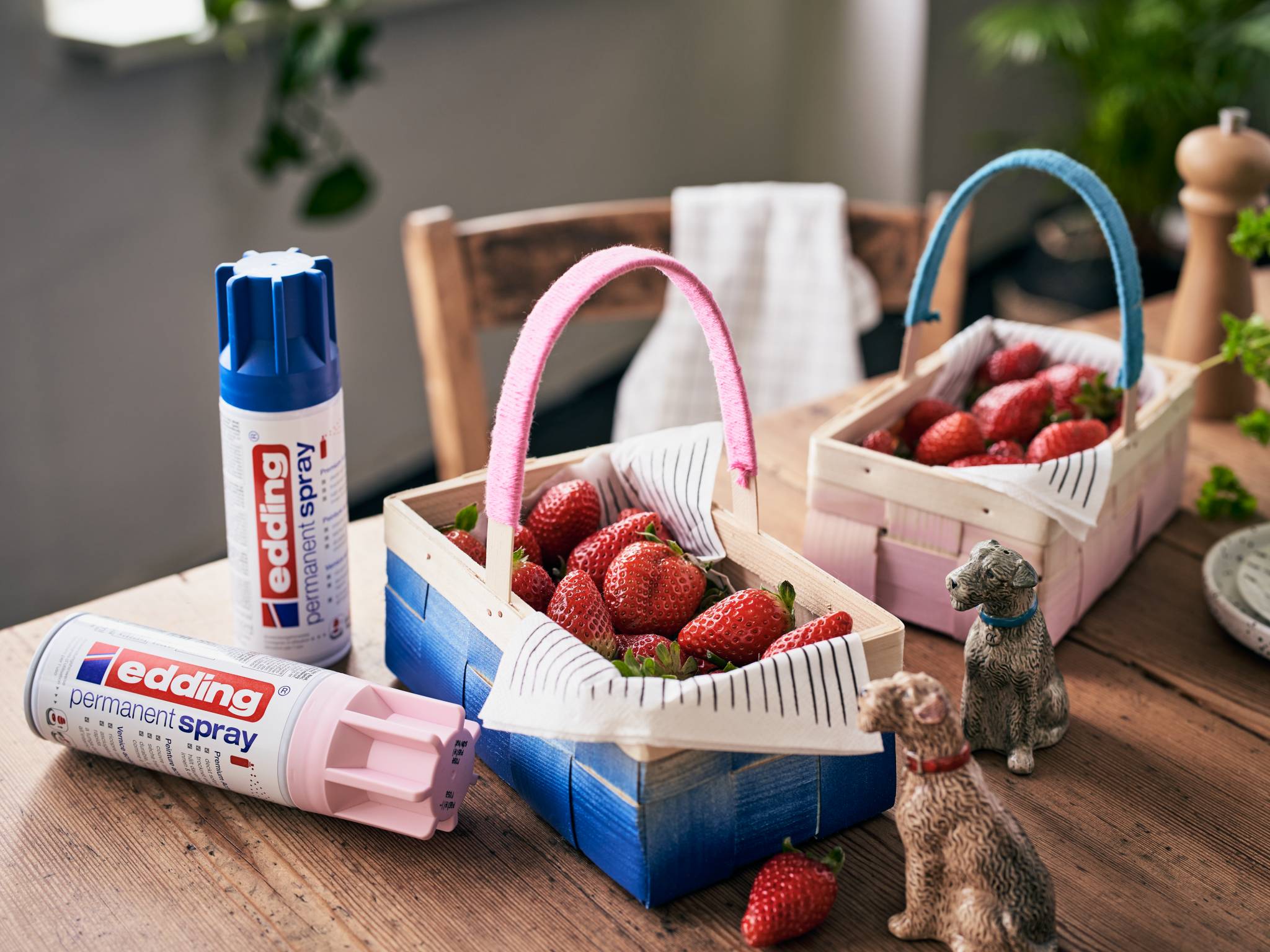 Zwei kleine Holzkörbe gefüllt mit Erdbeeren stehen auf einem Tisch neben zwei Edding-Spraydosen und kleinen Tierfiguren.