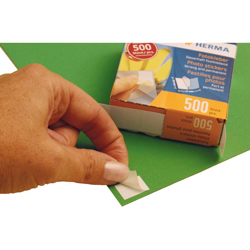 Eine Hand, die eine dreieckige Fotoecke auf ein grünes Blatt aufklebt. Eine Schachtel mit der Beschriftung 