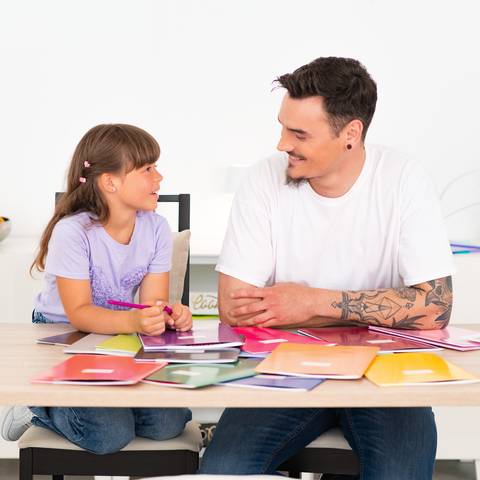 Mann und junges Mädchen sitzen an einem Tisch, lächeln und unterhalten sich, während sie auf bunte Notizbücher und Papiere schauen, die vor ihnen ausgebreitet sind.