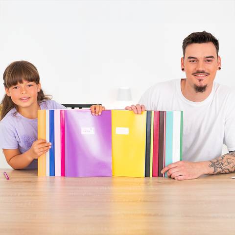 Ein lächelnder Mann und ein junges Mädchen sitzen an einem Tisch und halten bunte Ordner oder Notizbücher in Richtung der Kamera.
