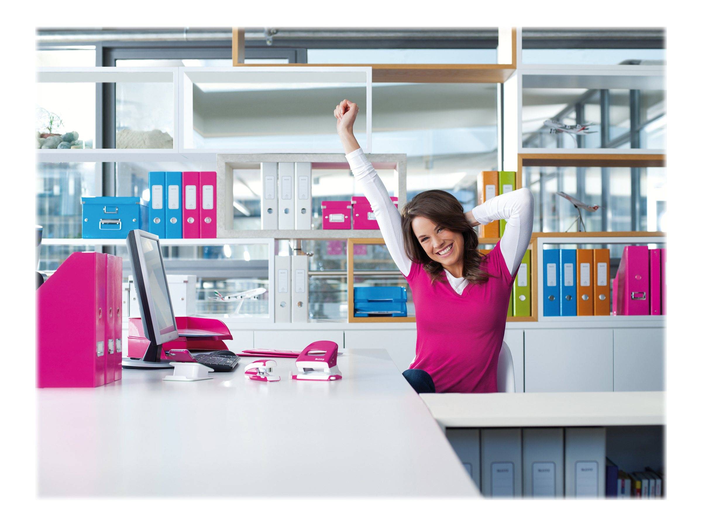Lächelnde Frau in einem rosa Top dehnt sich mit erhobenen Armen an einem Schreibtisch in einem hellen Büro, mit Computer, Telefon und farbigen Ordnern auf Regalen.