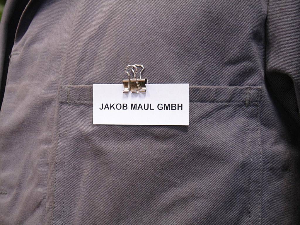 Ein Namensschild, das an einer Hemdtasche aus grauem Stoff angebracht ist, zeigt die Aufschrift 