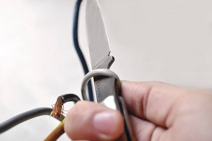Eine Hand verwendet ein Messer, um einen braunen Draht zu durchtrennen, wobei freiliegende Kupferstränge sichtbar werden, was einen Fokus auf elektrische Reparatur oder Verkabelungsarbeiten andeutet.
