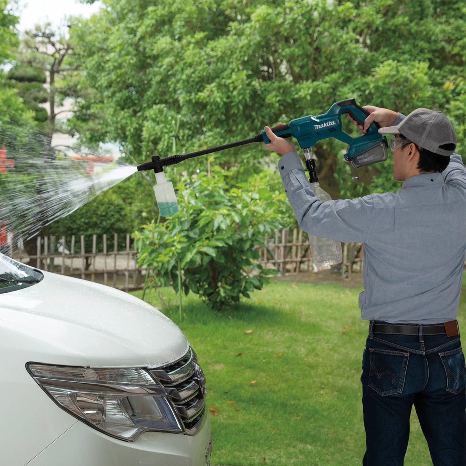 Eine Person verwendet einen Hochdruckreiniger, um die Windschutzscheibe eines weißen Autos in einer Gartenumgebung zu reinigen.