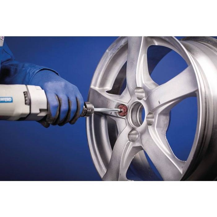 Eine Person in blauen Handschuhen verwendet einen Bohrer, um die Innenseite eines Silberlegierungsrads vor einem blauen Hintergrund zu polieren.