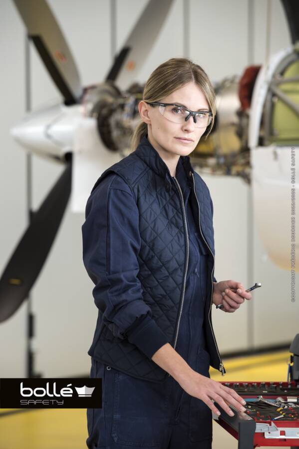 Weibliche Technikerin in Schutzbrille und dunkelblauer Arbeitskleidung steht neben einer Flugzeugschraube, hält ein Werkzeug und arbeitet an einem Teiletisch in einer Flugzeughalle.