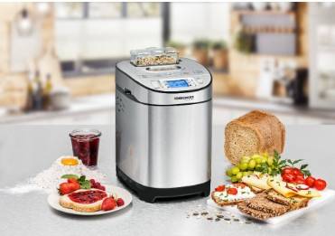Eine Brotbackmaschine aus Edelstahl auf einer Küchenarbeitsplatte, umgeben von einem Brotlaib, verstreuten Samen, frischen Früchten und einem Glas Saft.