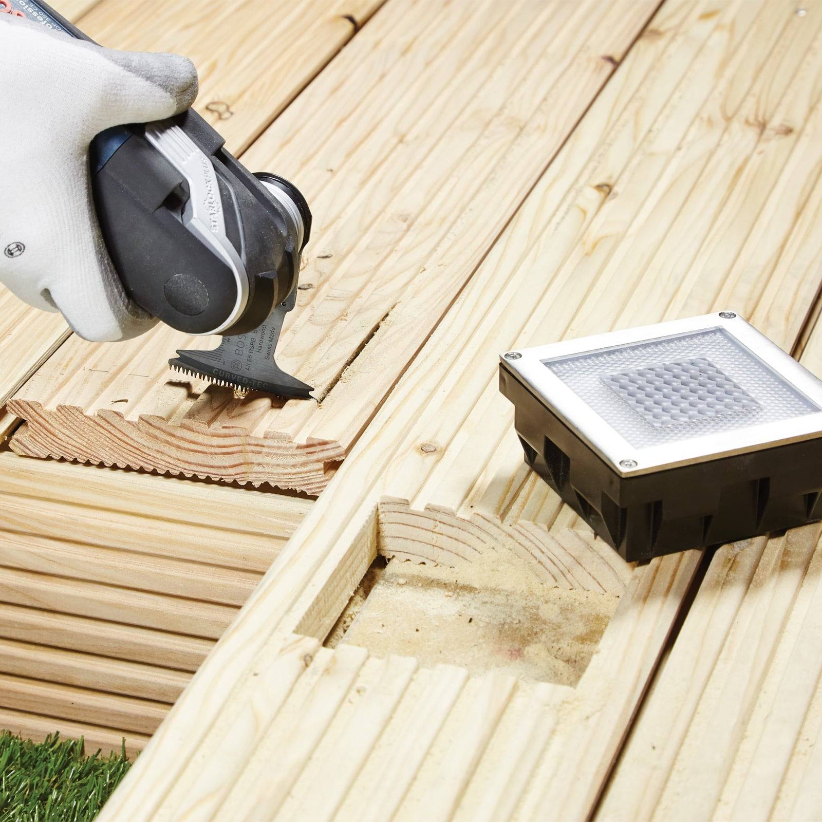 Eine Person, die ein Handwerkzeug verwendet, um eine Nut in einem Holzdeck neben einer eingelassenen Leuchte zu schneiden und eine Installation oder Reparatur vorbereitet.