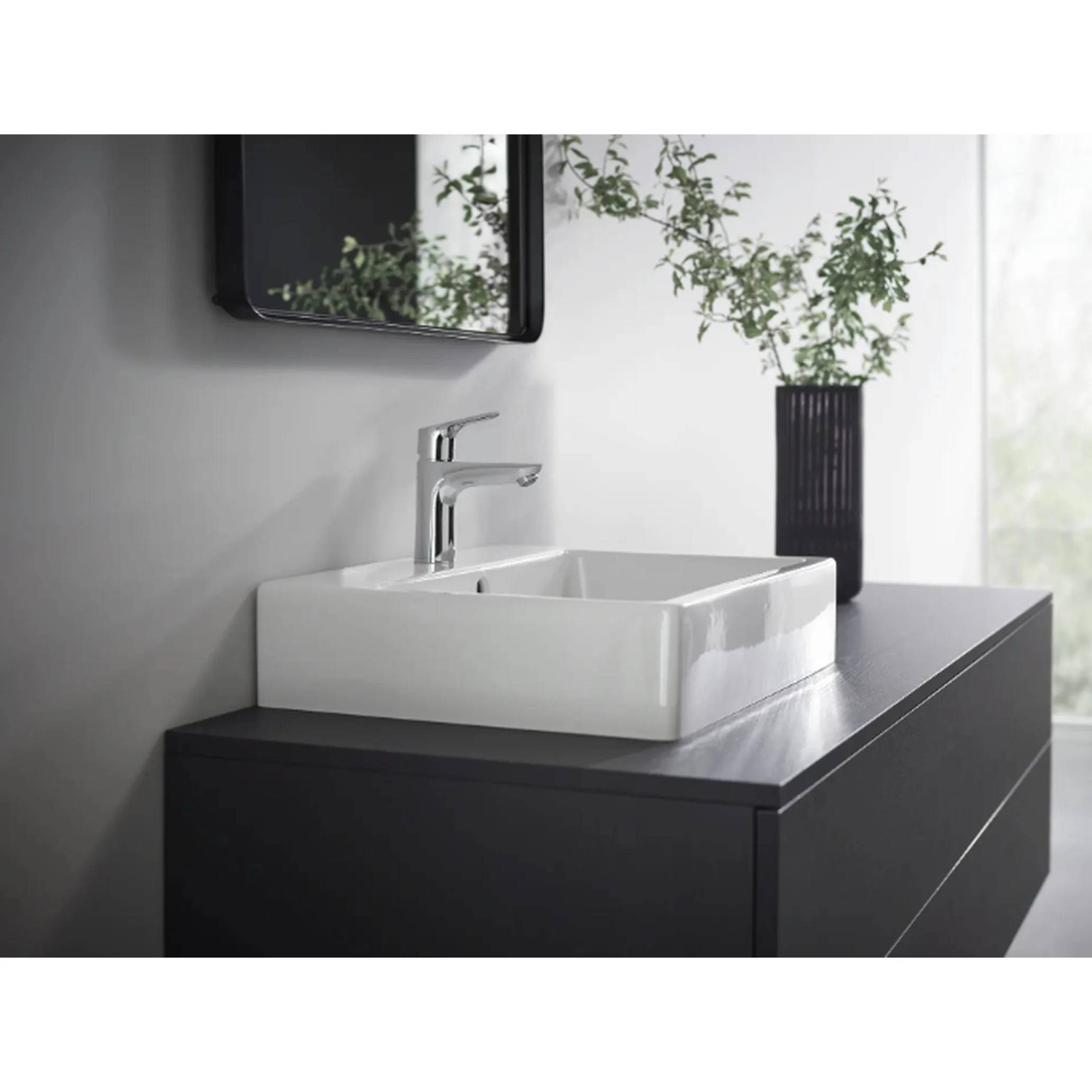 Modernes Badezimmer-Waschtisch mit einem weißen quadratischen Aufsatzwaschbecken und chromfarbener Armatur auf einem dunklen Schrank, daneben eine Vase mit Grünpflanzen.