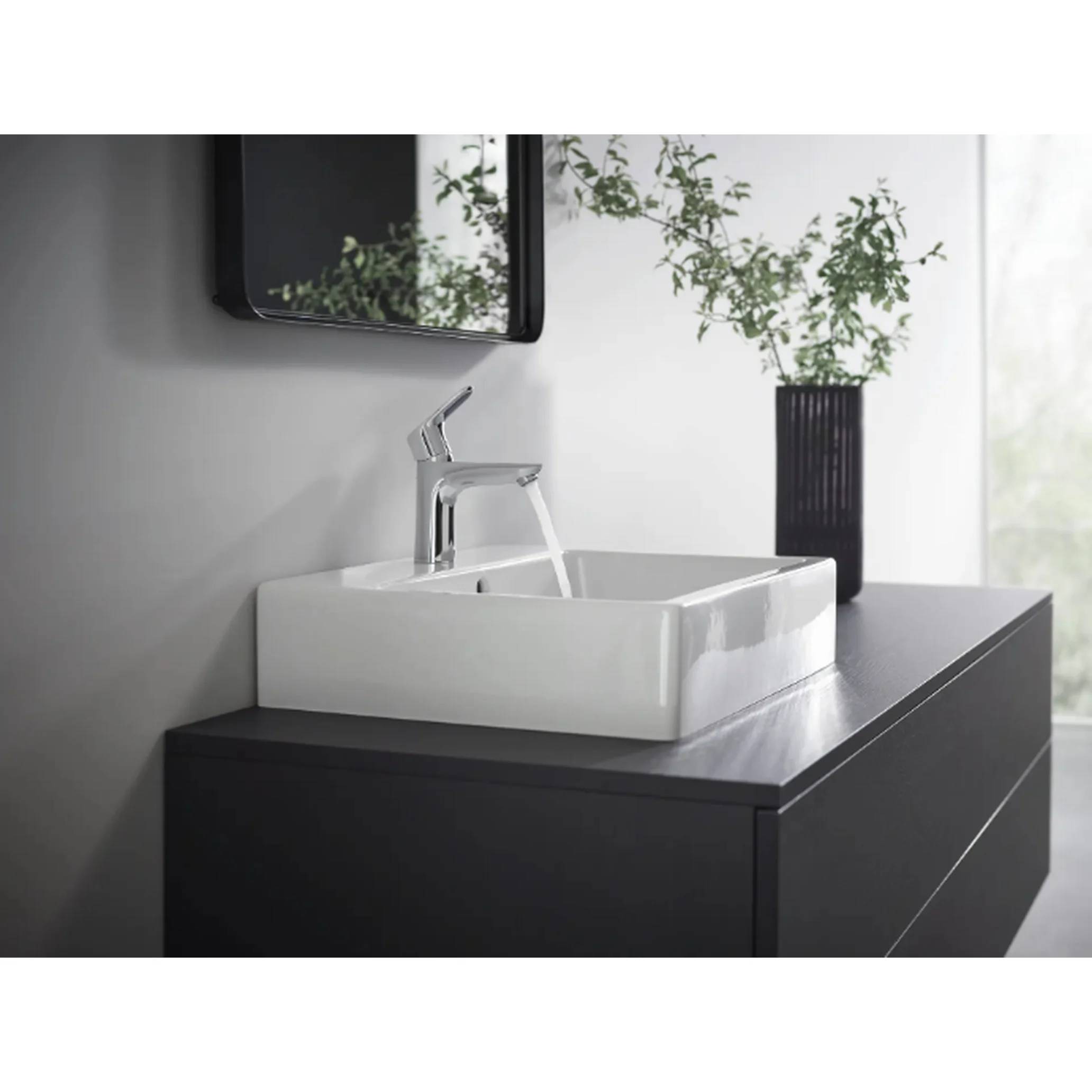 Modernes Badezimmerbecken mit Chromarmatur auf einer dunklen Waschtischanlage, einem schwarzgerahmten Spiegel und einer Vase mit Grünpflanzen vor einer hellen Wand.