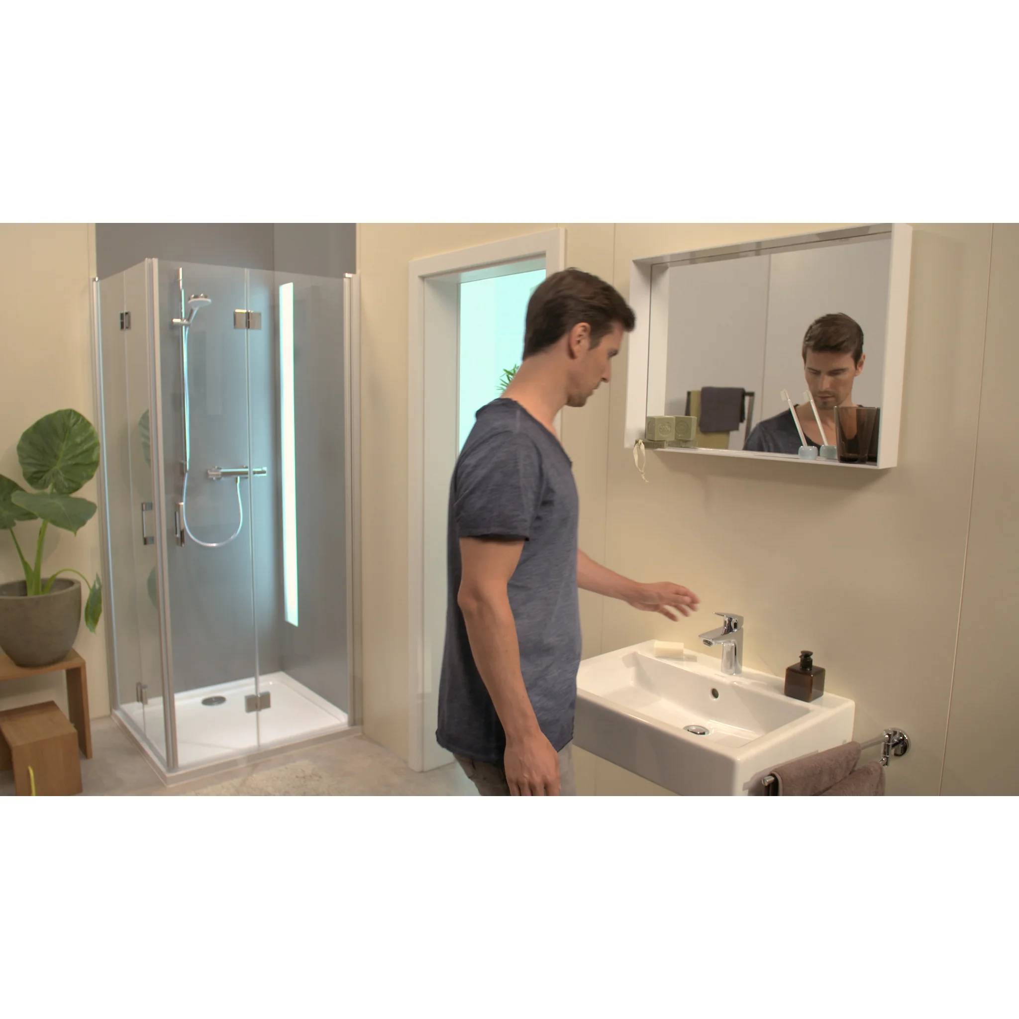 Ein Mann im Badezimmer steht an einem Waschbecken und schaut in den Spiegel, mit einer Duschkabine und einer Topfpflanze im Hintergrund.