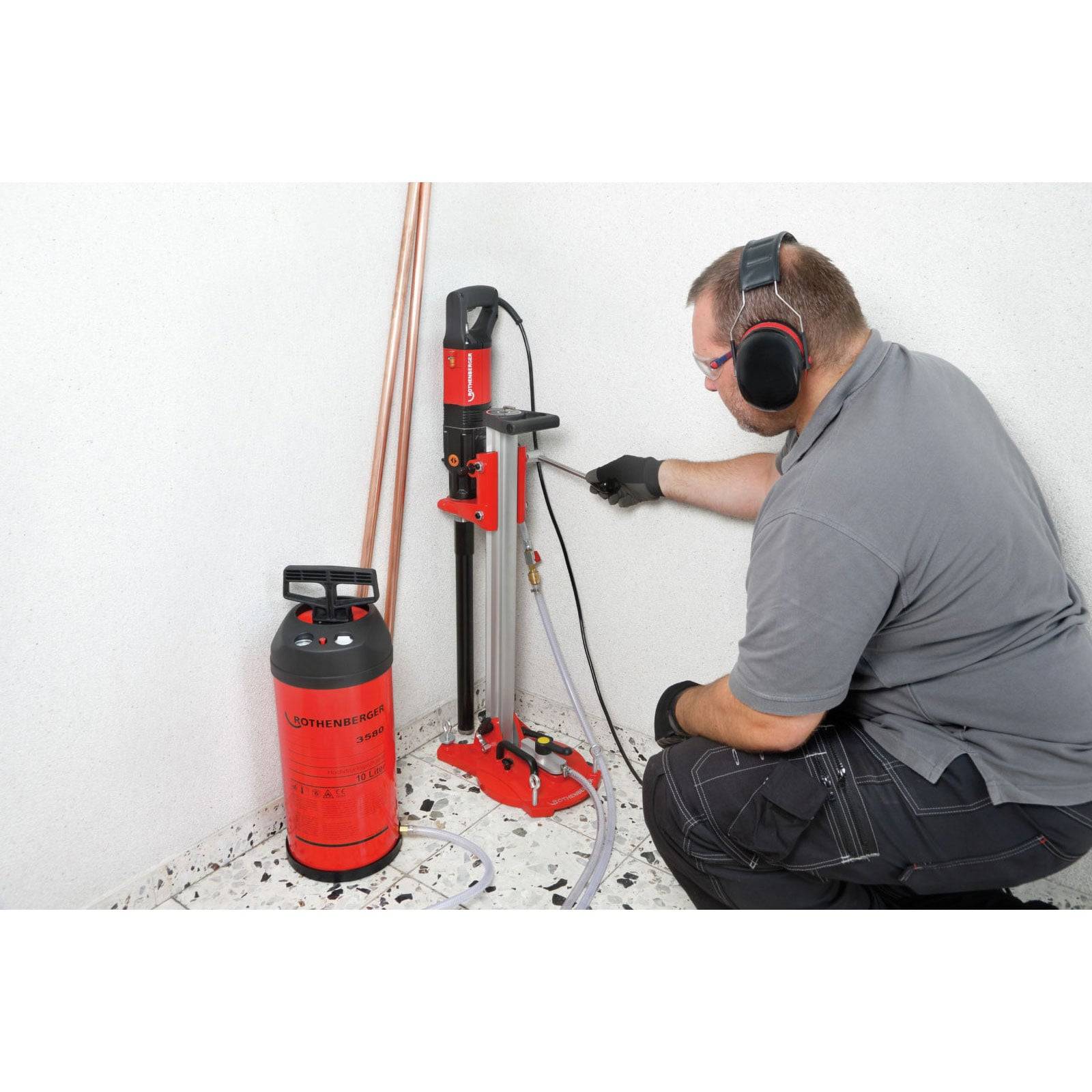 Eine Person, die ein Kernbohrgerät mit Gehörschutz verwendet. Ein roter Feuerlöscher und Kupferrohre liegen zur Sicherheit auf dem Boden.