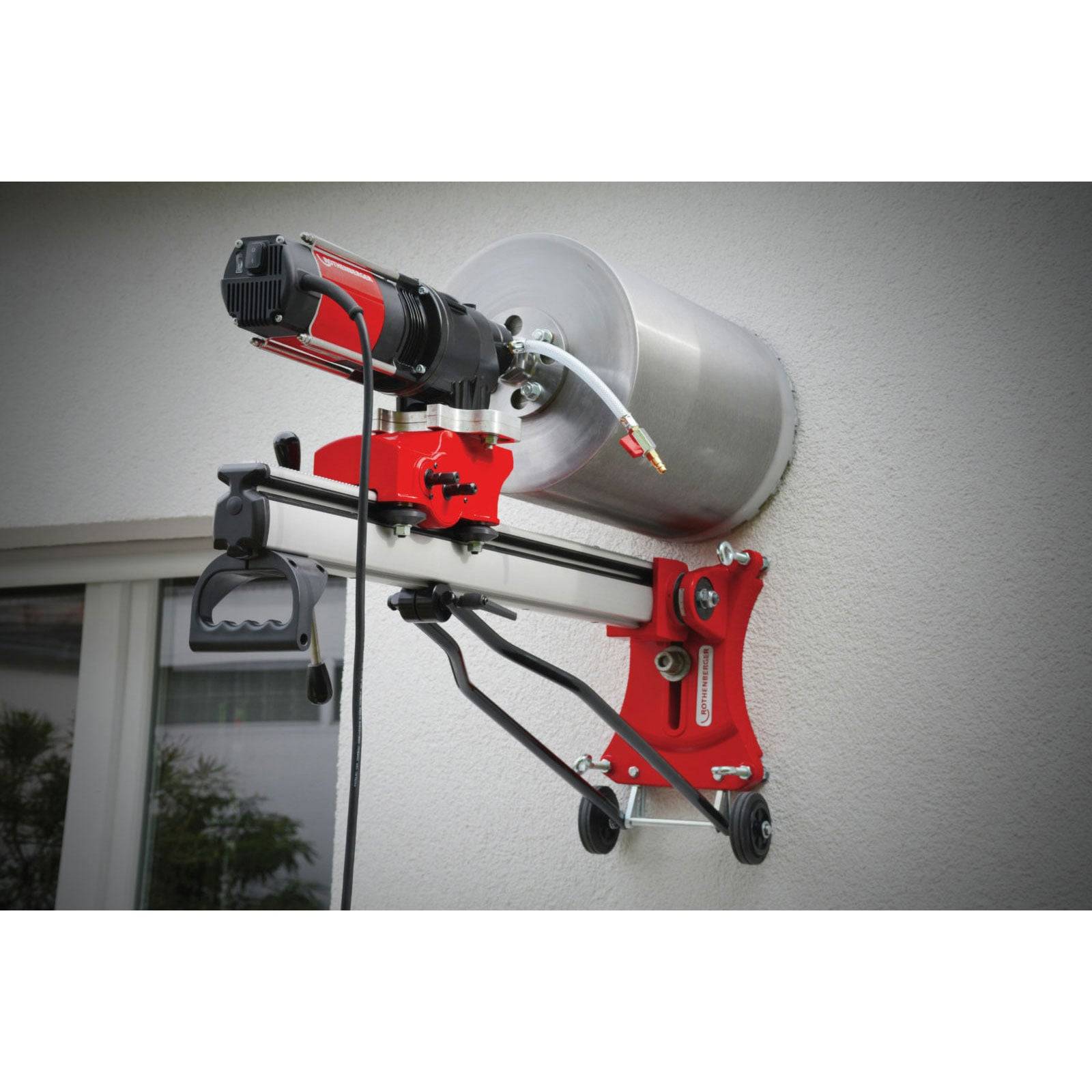 Ein rot-silberner, wandmontierter zylindrischer Apparat mit Griff und Kabeln, möglicherweise ein Elektrowerkzeug, ist an der Außenwand eines Gebäudes befestigt.