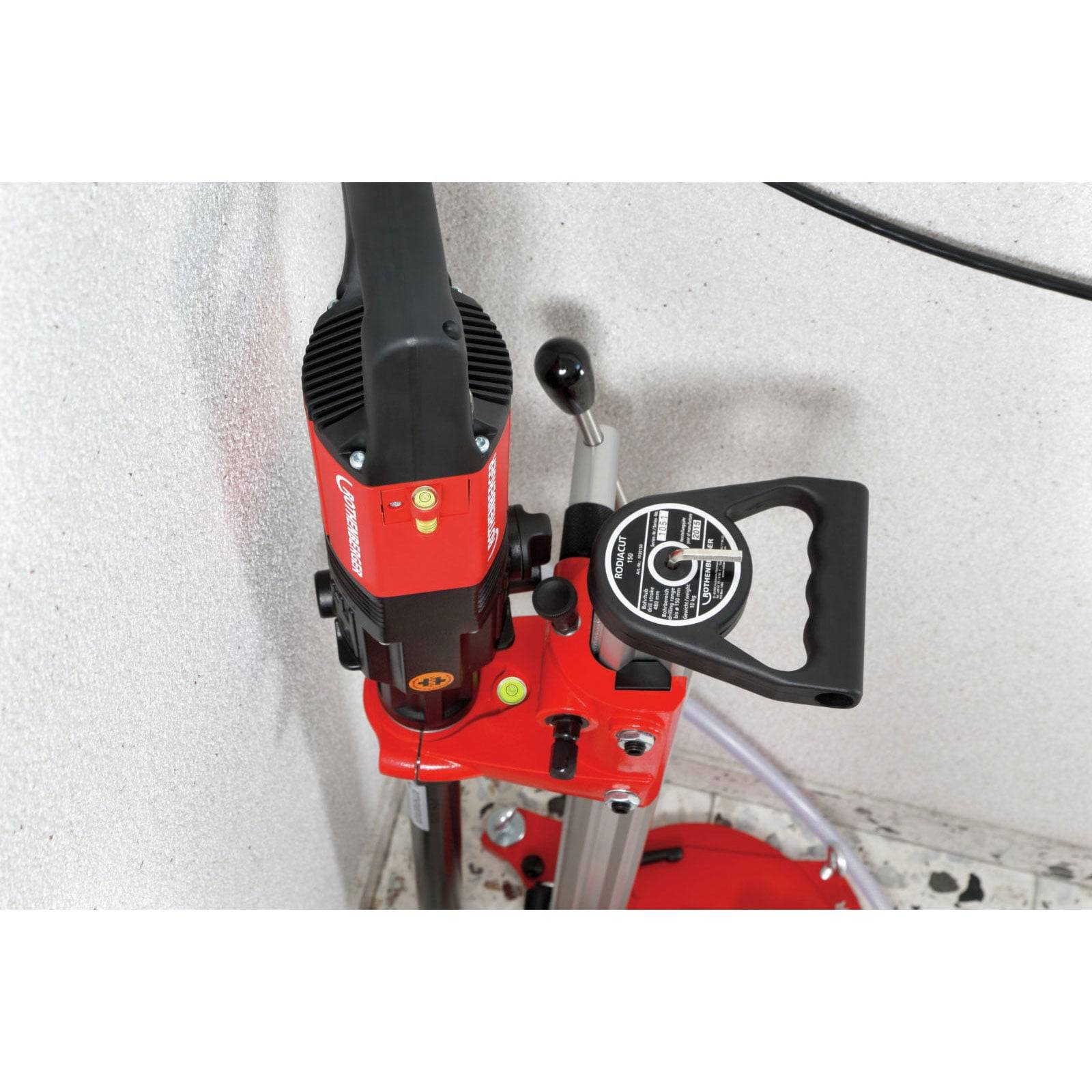 Ein roter Bohrer, montiert auf einem stationären Ständer, in einer Ecke an einer weißen Wand positioniert. Der Bohrer hat einen sichtbaren Bedienungsgriff und ein schwarzes Netzkabel.