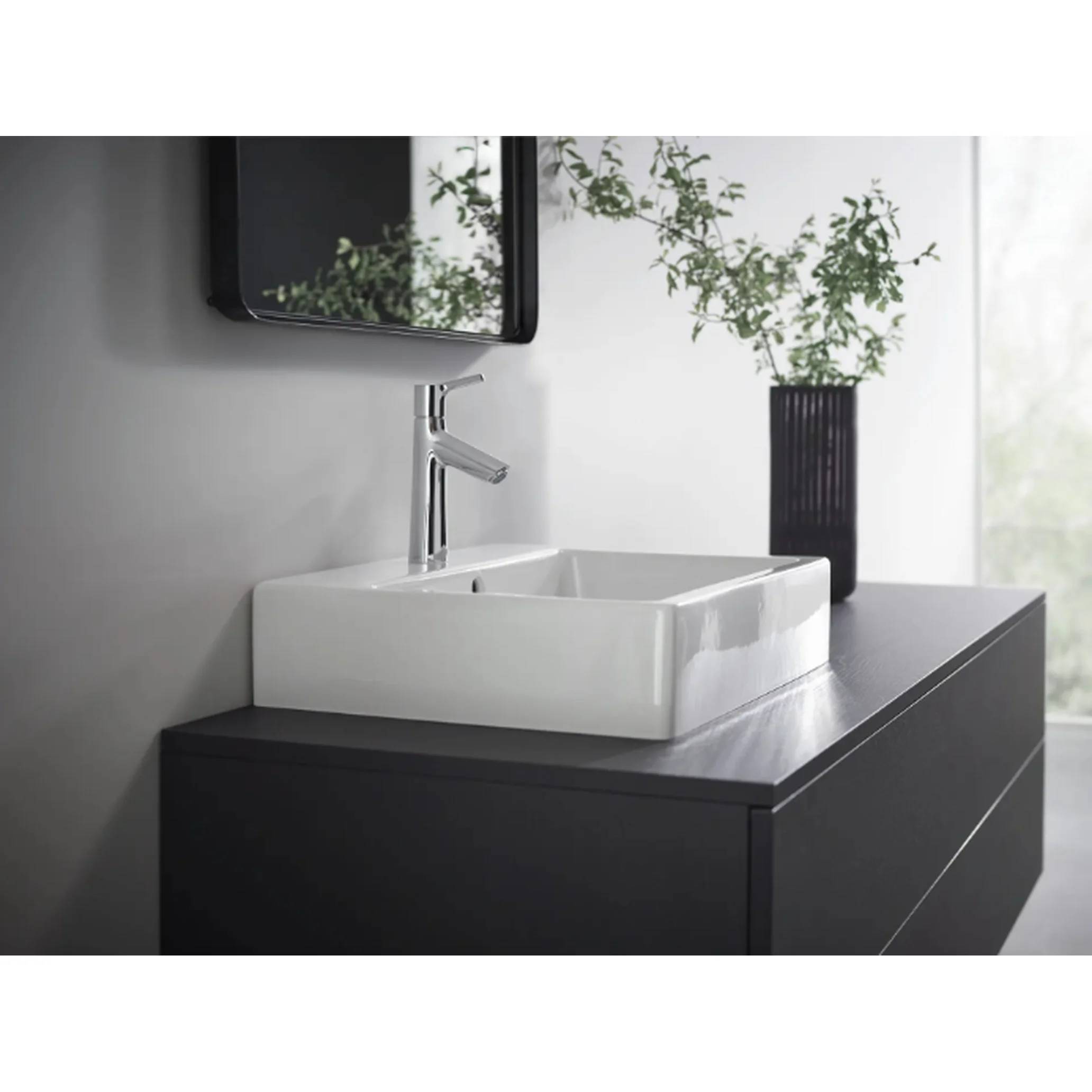 Modernes Badezimmer-Waschtisch mit weißem quadratischem Aufsatzwaschbecken, Chromarmatur, Wandspiegel und schwarzem Schrank neben einer Vase mit Grünpflanzen.