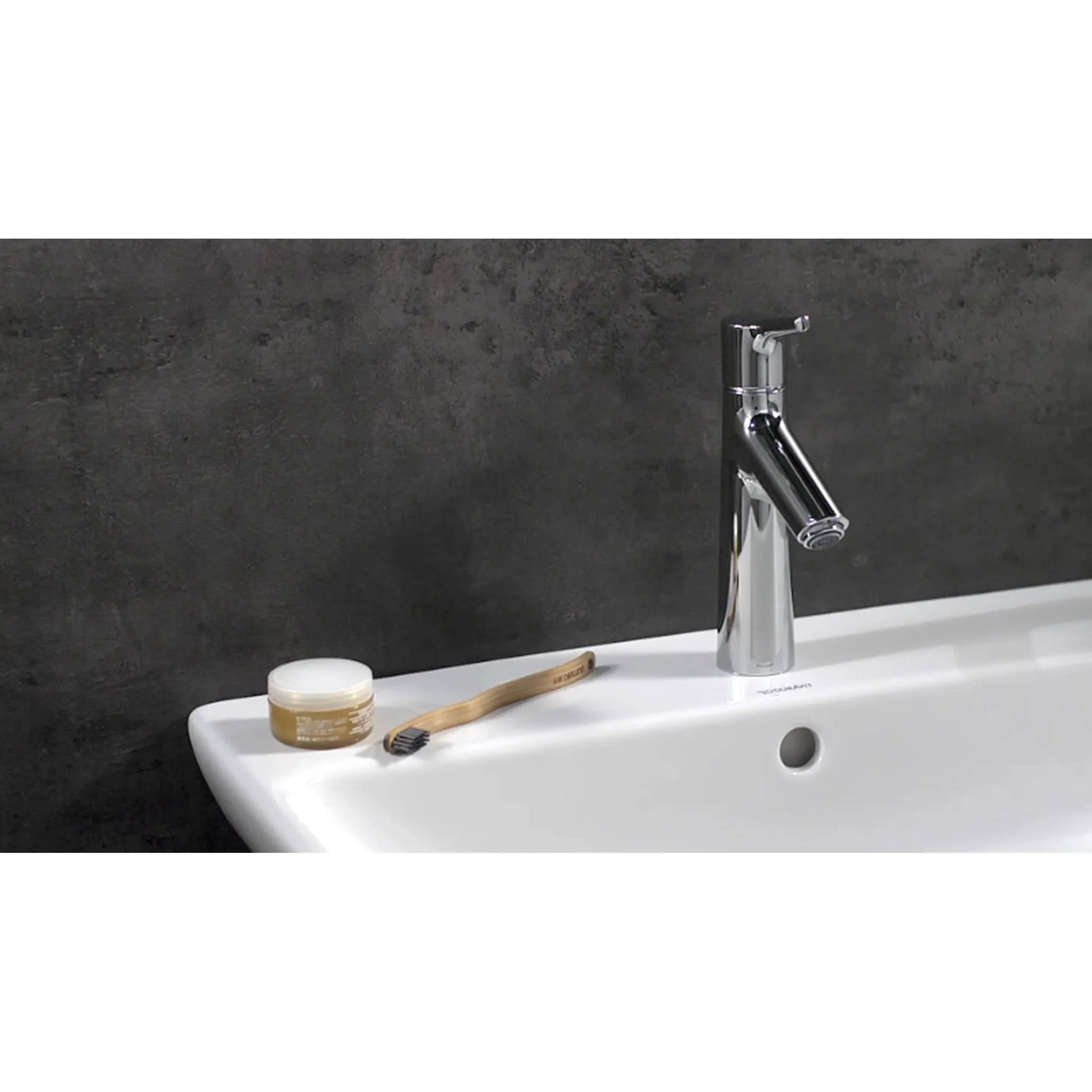 Modernes Badezimmerbecken mit Chromarmatur, Seifenglas und Zahnbürste auf einem weißen Becken vor einer dunkel strukturierten Wand.