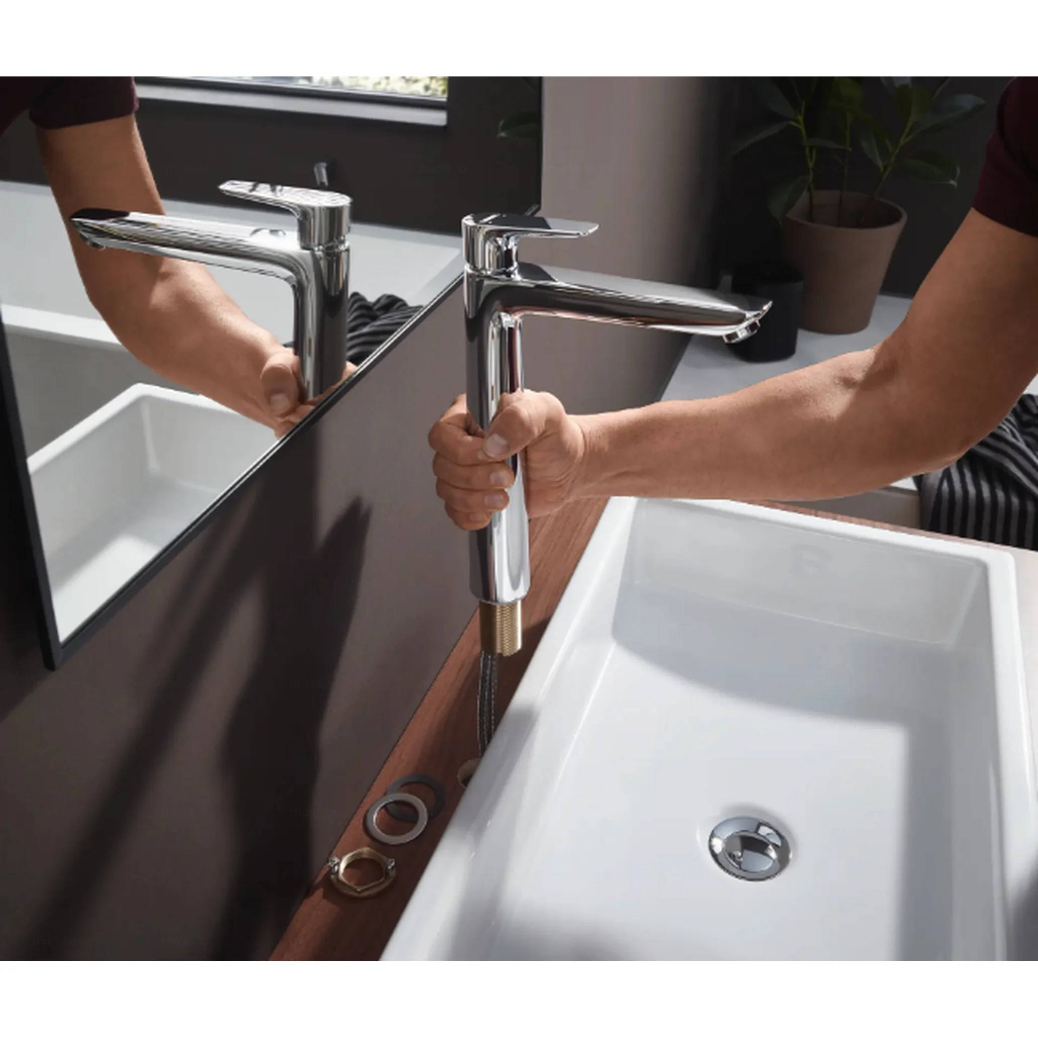 Person installiert einen verchromten Badezimmerhahn auf einem weißen Waschbecken neben einem Spiegel und zeigt die Sanitärinstallation, die eingebaut wird.