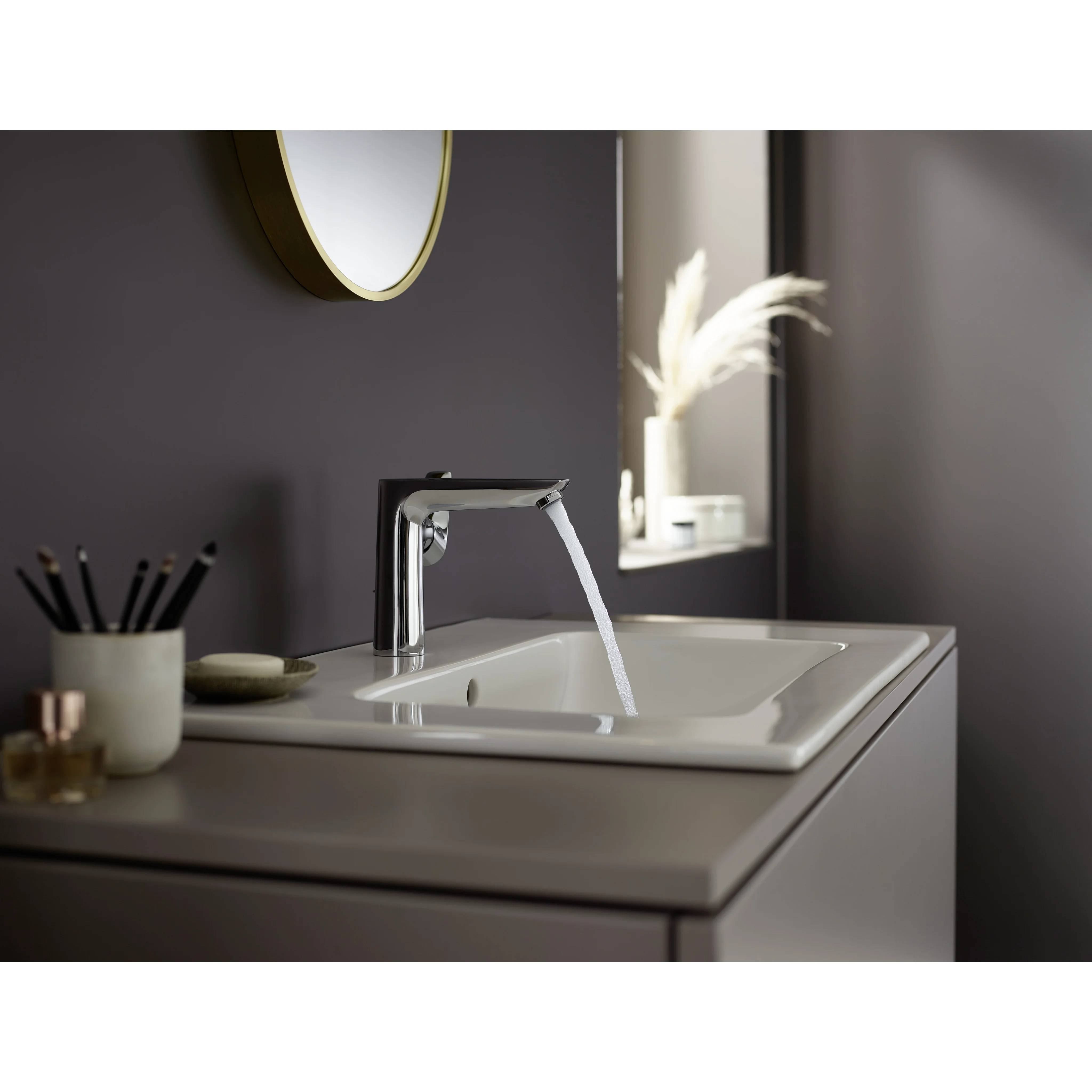 Modernes Badezimmerbecken mit Chromarmatur, aus der Wasser in ein weißes Becken fließt, neben einem Spiegel und kleinen Toilettenartikeln auf der Arbeitsplatte.