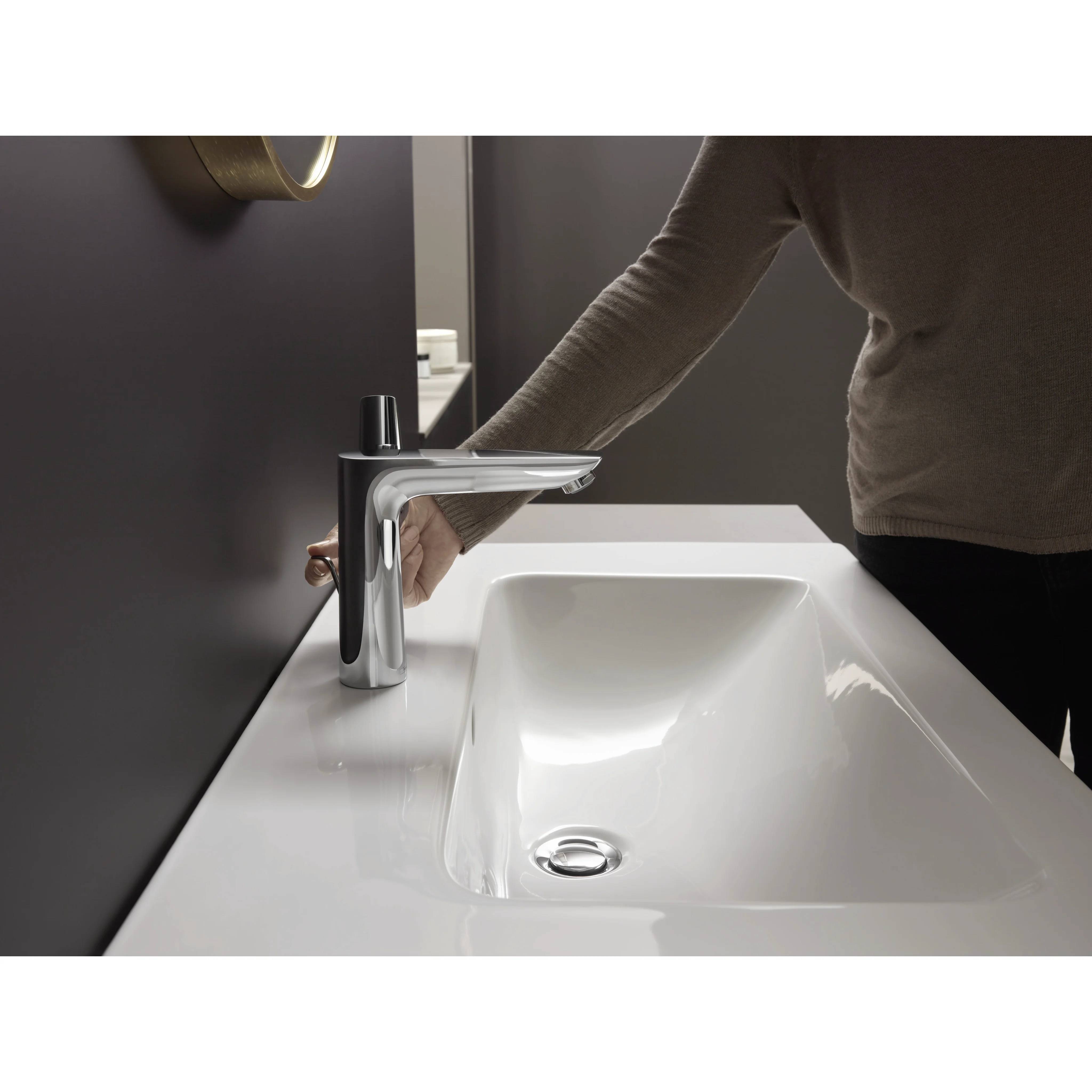Eine Person greift nach einem modernen Chrom-Badezimmerhahn neben einem weißen Waschbecken in einem minimalistischen Badezimmer.