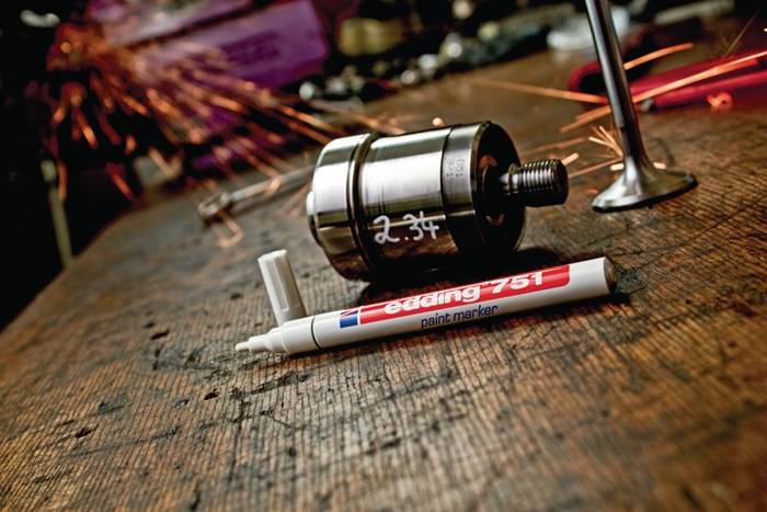 Ein Metallobjekt mit der Beschriftung 2,34 liegt auf einer Werkbank, neben dem ein Edding 751 Lackmarker platziert ist, während im Hintergrund Funken sprühen.