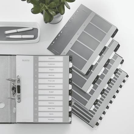 Stapel grauer und schwarzer Ordnertrenner mit nummerierten Reitern und Beschriftungsfeldern auf einem Schreibtisch neben Stiften, was auf Büroorganisation hindeutet.