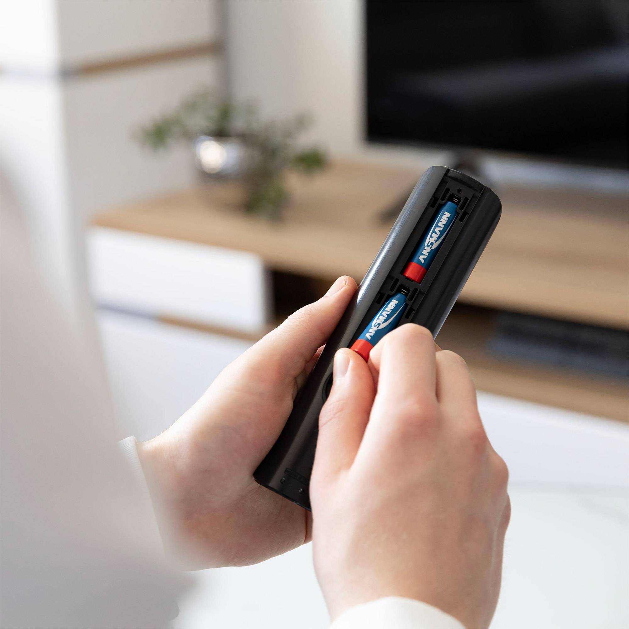 Hände führen Batterien in eine schwarze Fernbedienung in einem Wohnzimmer ein, mit einem verschwommenen Fernseher und Schrank im Hintergrund.