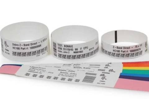 Drei weiße Identifikationsbänder mit gedruckten Patientendetails und Barcodes, darunter ein regenbogenfarbenes Musterblatt.