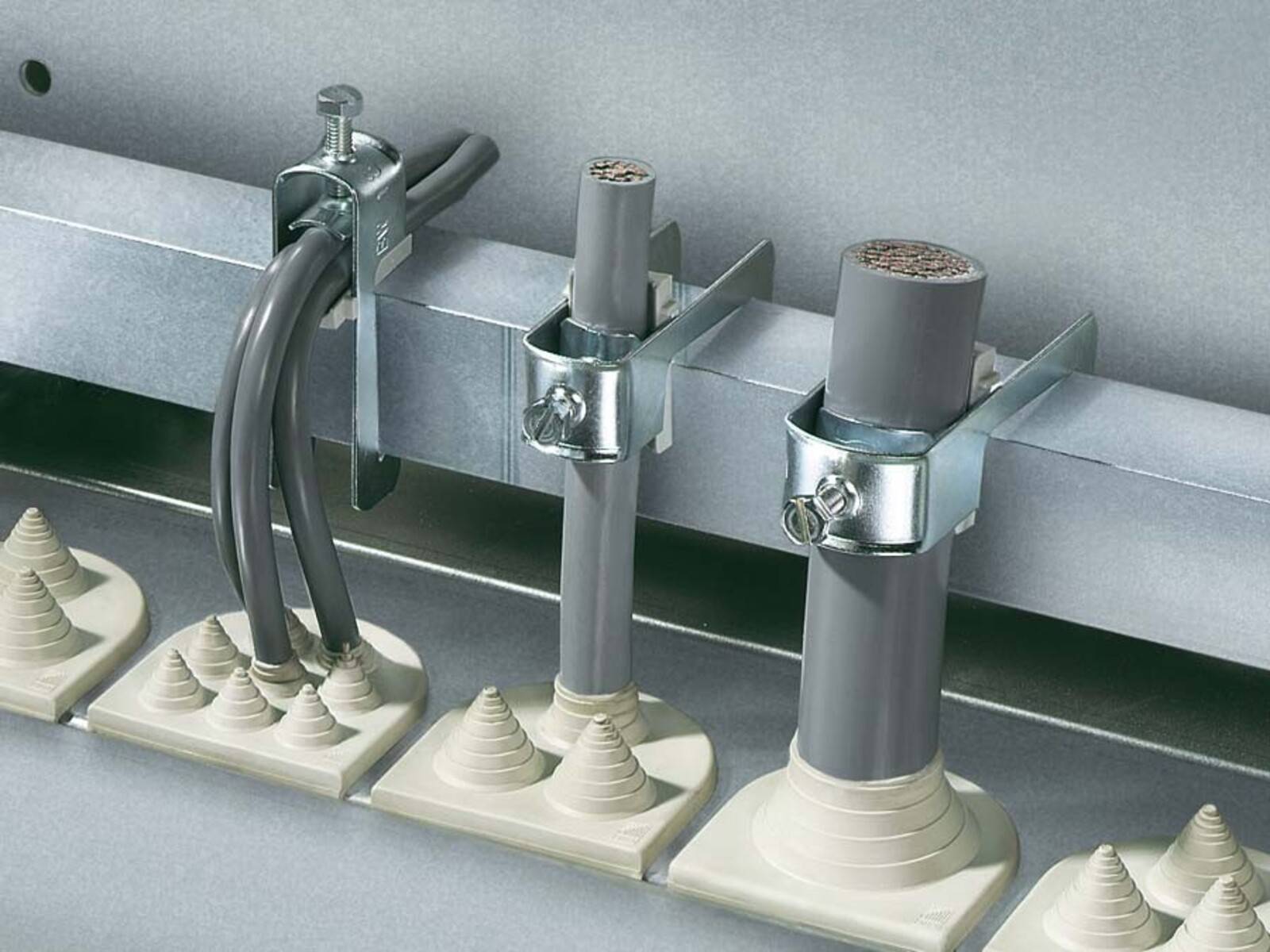 Zwei graue Kabel sind mit Metallklemmen befestigt und führen in senkrechte graue Rohrleitungen. Der Aufbau deutet auf Kabelmanagement für elektrische Systeme hin.