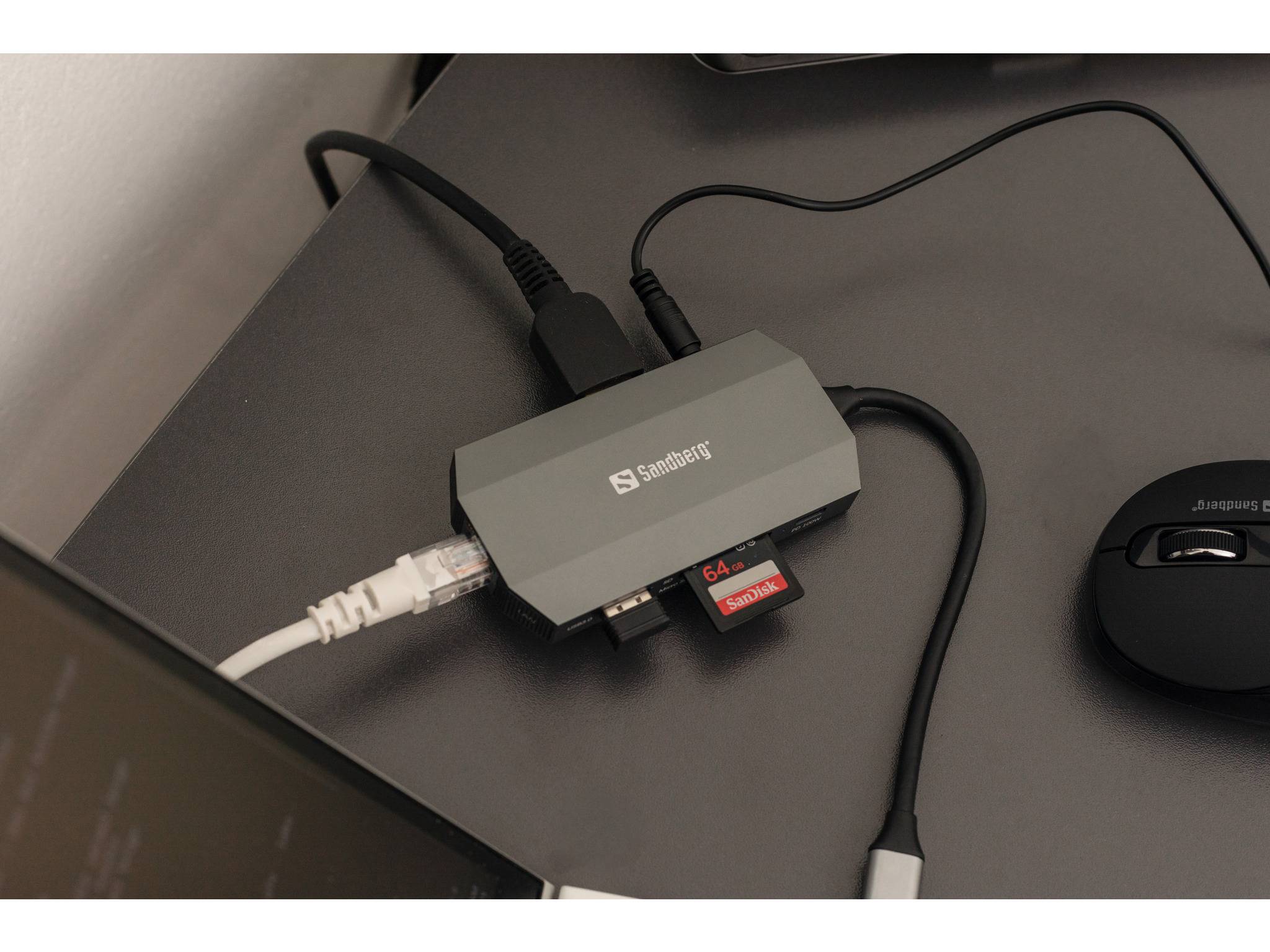 Ein Sandberg USB-Hub auf einem Schreibtisch verbindet ein Ethernet-Kabel, eine Speicherkarte und andere Peripheriegeräte, mit einer Maus in der Nähe.