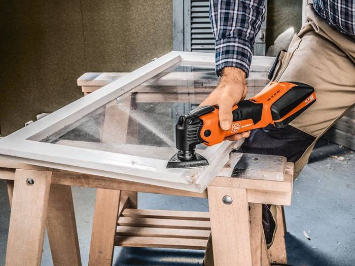Eine Person verwendet eine elektrische Schleifmaschine, um die Kante eines Holzrahmens zu glätten, der auf einer Werkbank liegt und damit Holzbearbeitung in Gange ist.