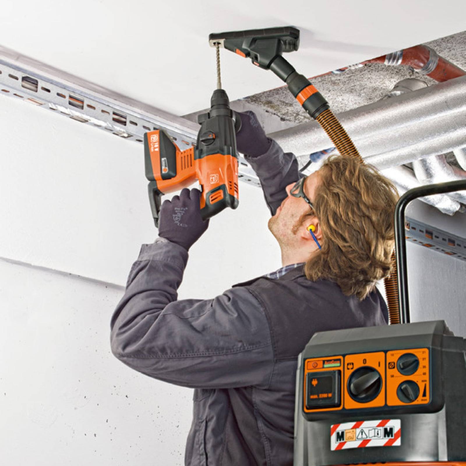 Ein Arbeiter verwendet einen Bohrer, um einen Bolzen in eine Stahldeckenstütze einzuschrauben, dabei trägt er Sicherheitsbrille und Handschuhe. Ein Kontrollpanel mit Drehknöpfen ist zu sehen.