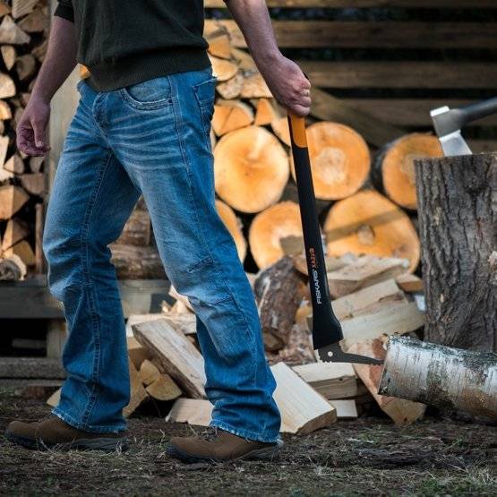 Eine Person in Jeans hackt Holz mit einer Axt und ist von Stämmen und Brennholz umgeben, was auf eine Holzfälleraktivität im Freien hindeutet.