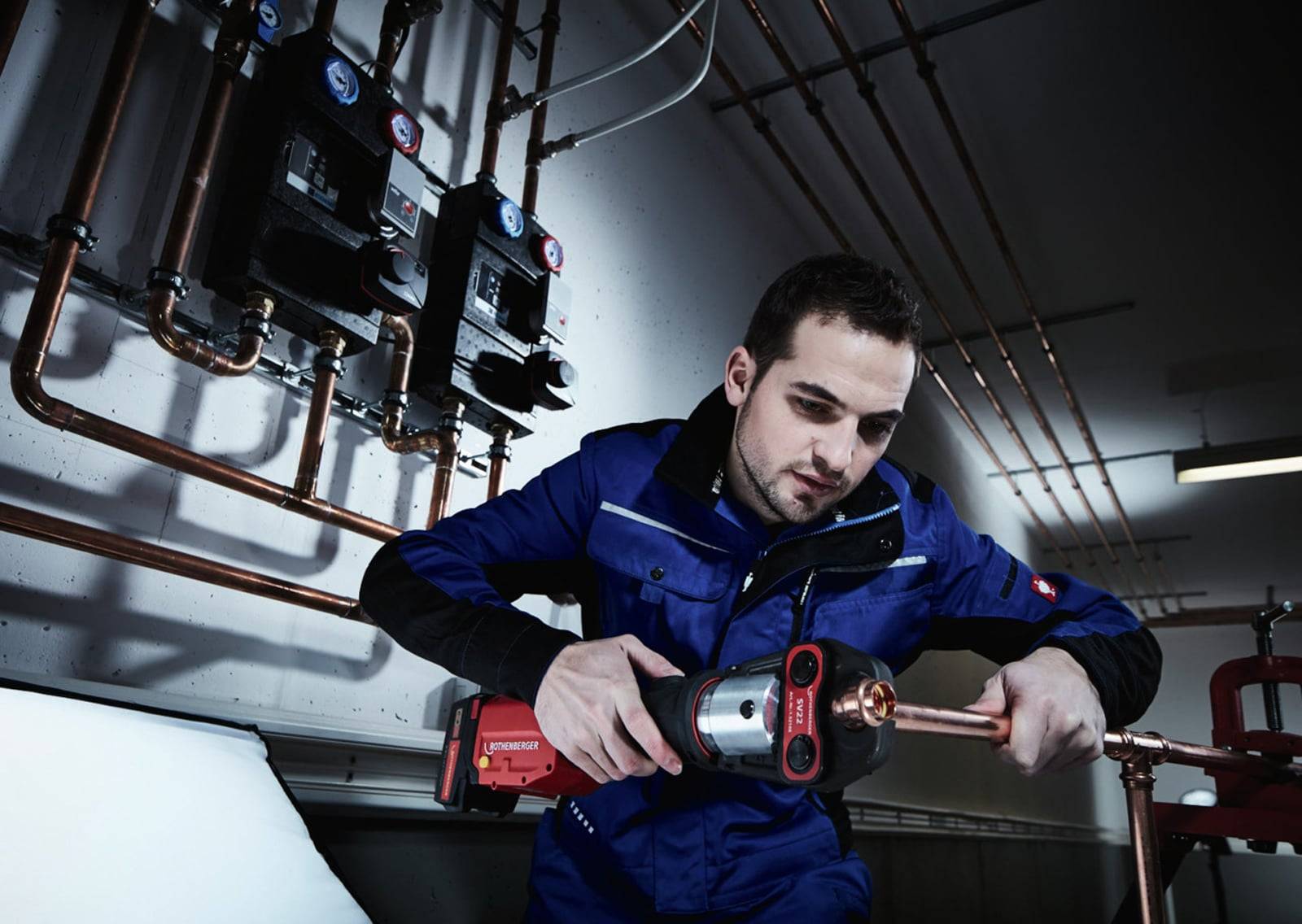 Eine Person in blauer Uniform bedient ein hydraulisches Werkzeug an Kupferrohren, die Teil einer Sanitär- oder Heizungsanlage sind, mit Messgeräten im Hintergrund.