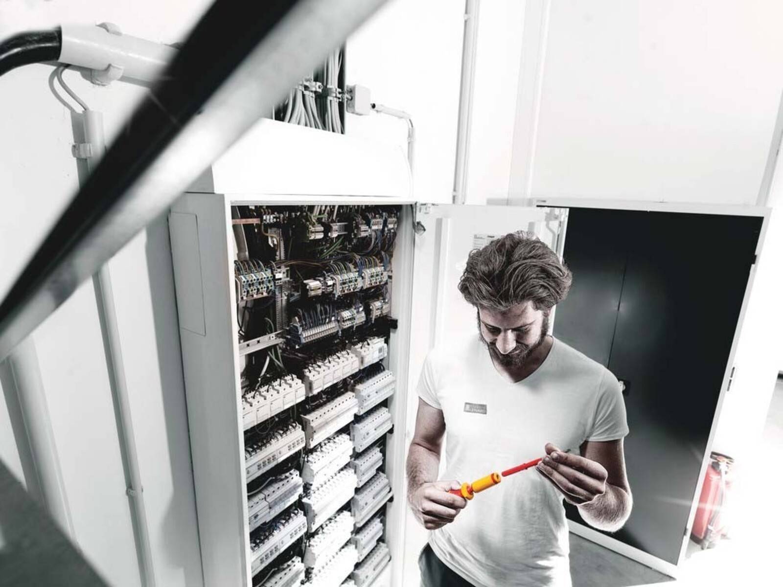 Ein Mann in einem weißen Hemd arbeitet an einer elektrischen Tafel und untersucht Kabel mit einem Schraubendreher, wobei er sich auf die Verbindungen im Inneren konzentriert.