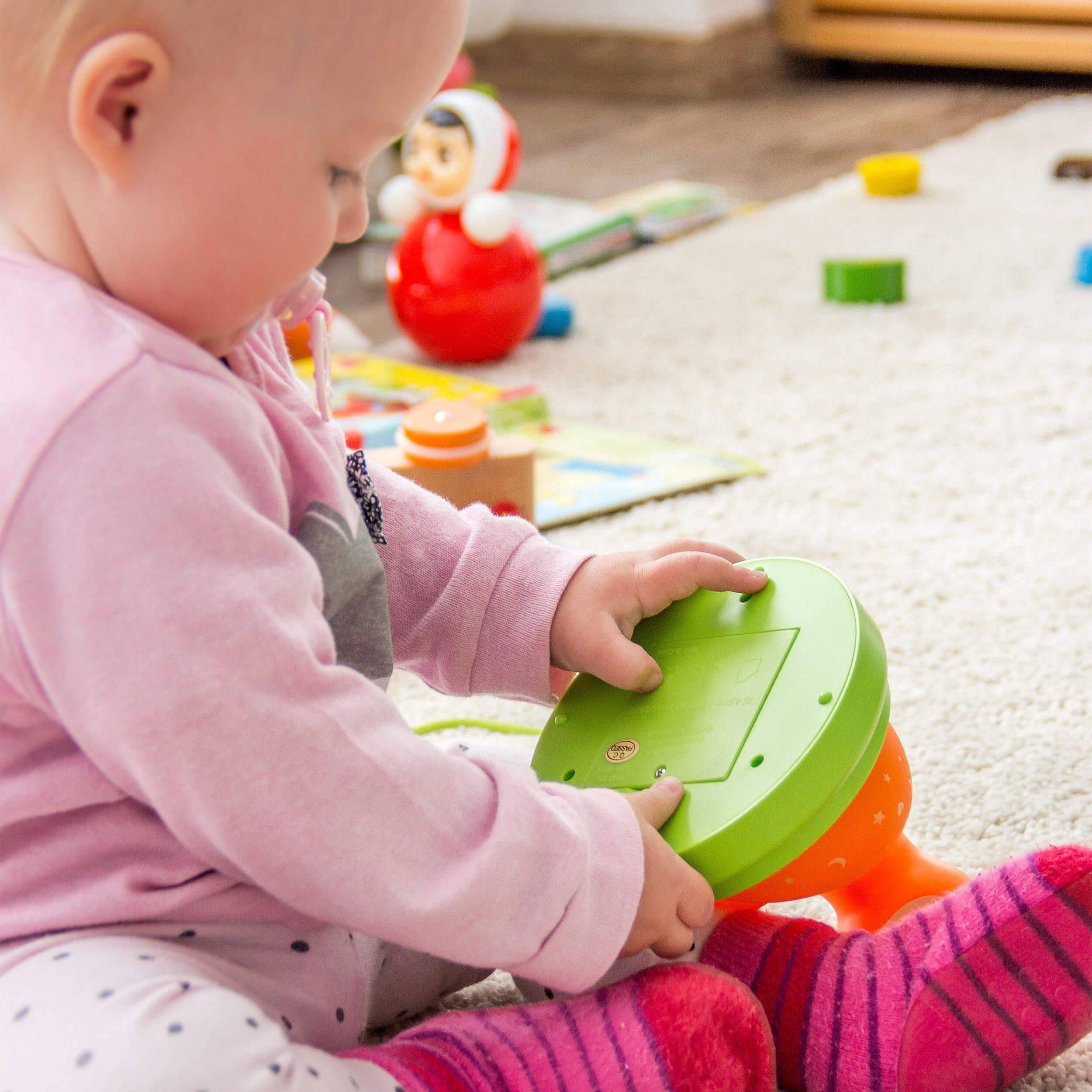 Baby in rosa Hemd spielt mit einem grün-orange Spielzeug auf einem Teppichboden, mit anderen Spielzeugen verstreut im Hintergrund.