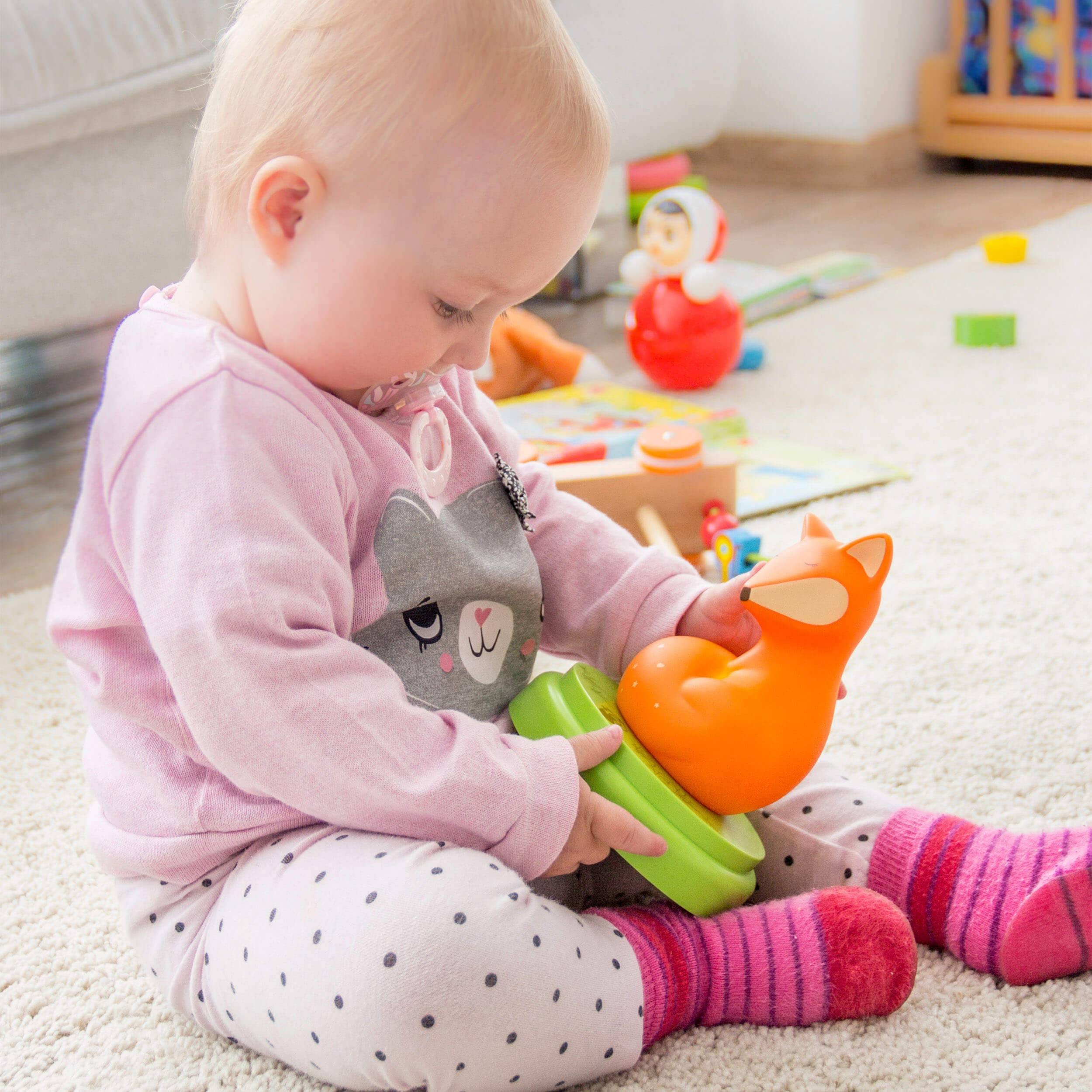 Ein Baby sitzt auf dem Boden und spielt mit einem orangefarbenen Spielzeugfuchs, konzentriert darauf, farbige Ringe in einem hellen Kinderzimmer zu stapeln.