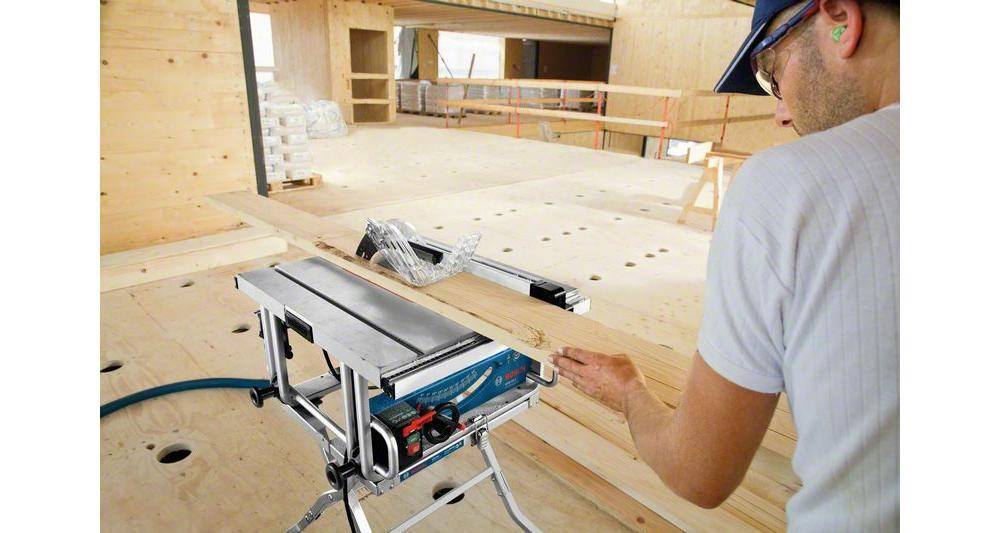 Eine Person verwendet eine Tischkreissäge, um ein Holzstück auf einer Baustelle zu sägen. Der Bereich befindet sich im Bau mit sichtbarem Holz und Werkzeugen.