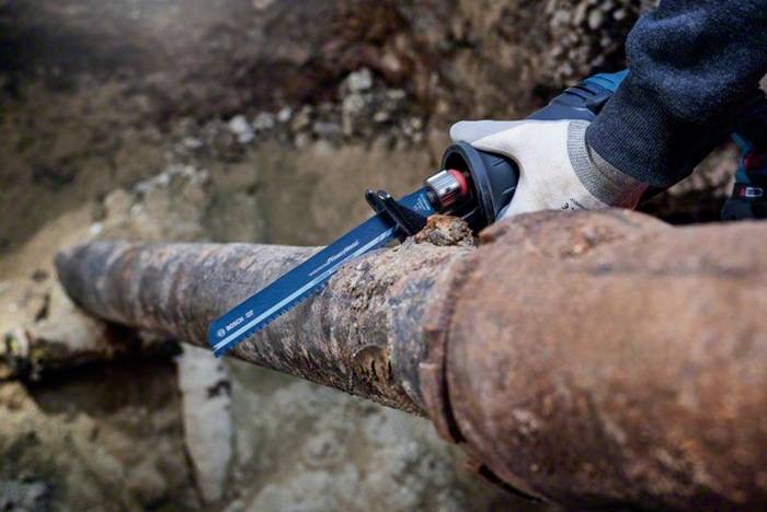 Eine Säbelsäge schneidet durch ein großes, verrostetes Metallrohr. Die Sägeblatt ist sichtbar in das Rohr einschneidend, von einer behandschuhten Hand ruhig gehalten.