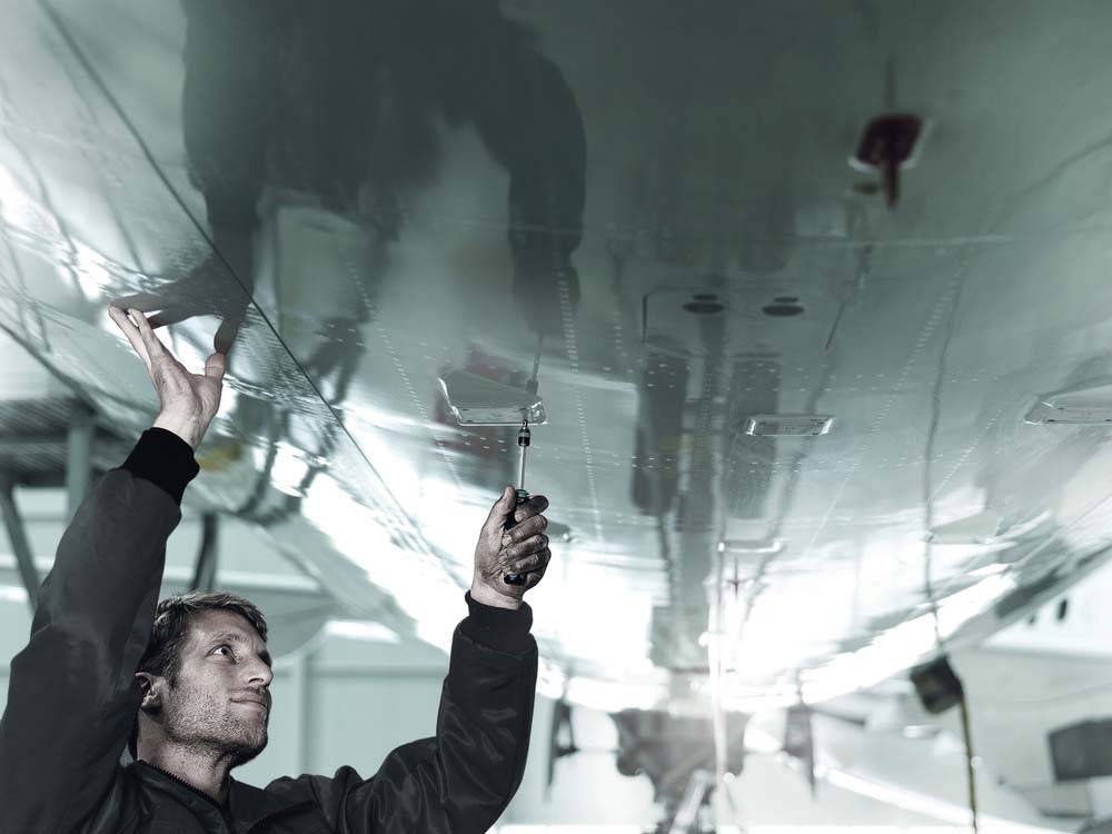 Eine Person, die unter dem Flügel eines Flugzeugs in einer Wartungshalle arbeitet und ein Werkzeug nach oben in Richtung der Flügeloberfläche hält, konzentriert auf ihre Aufgabe.