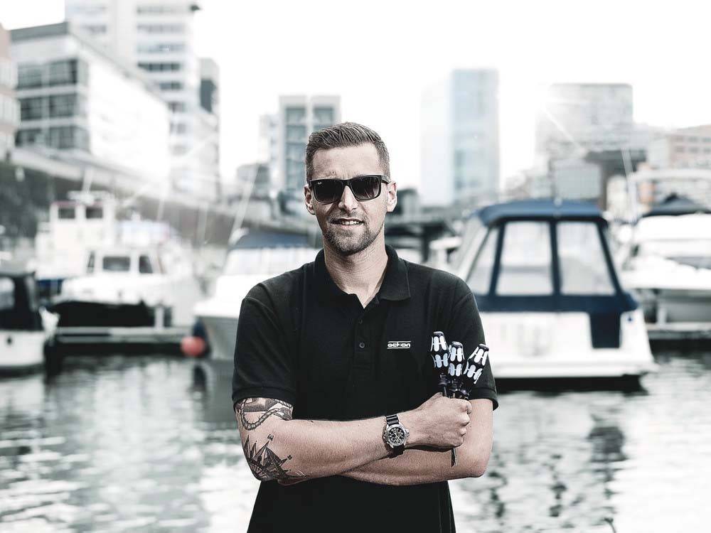 Eine Person mit Sonnenbrille und Tattoos steht mit verschränkten Armen vor einer Marina mit Booten und Stadtgebäuden im Hintergrund.