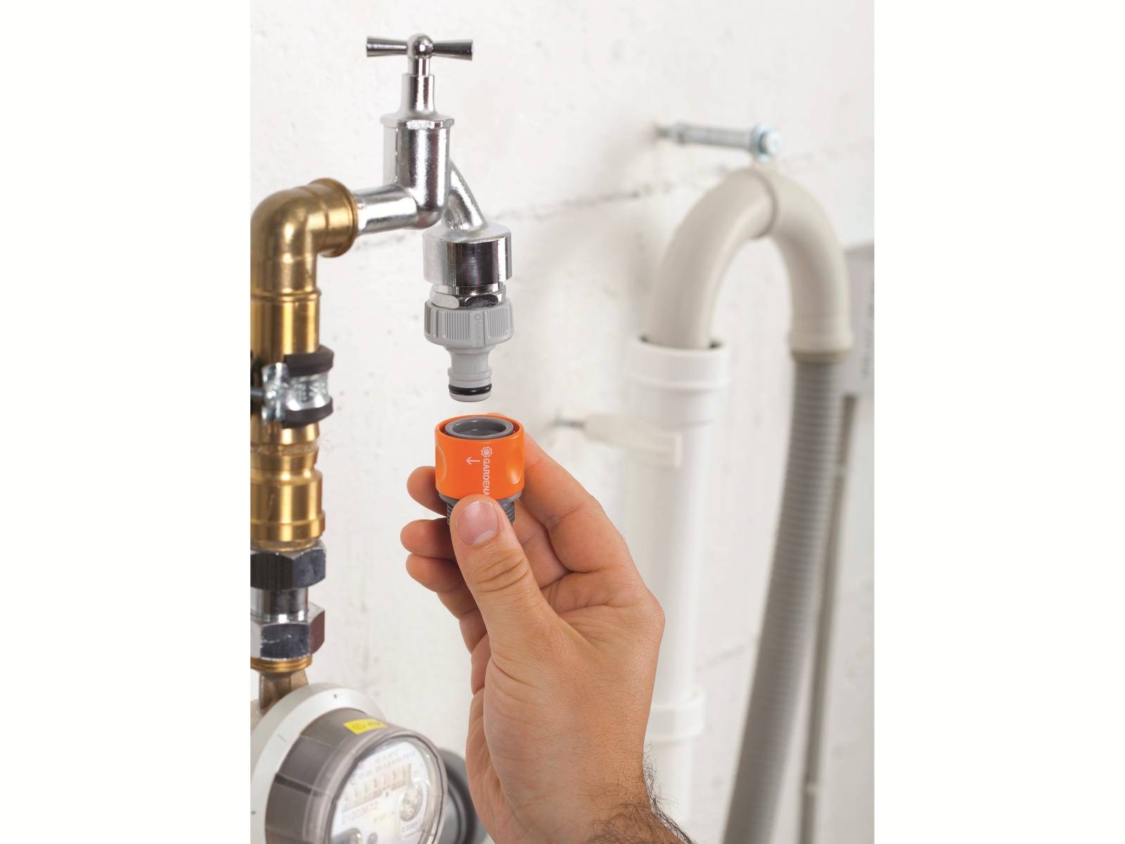 Eine Hand verbindet eine orangefarbene Schlauchkupplung mit einem metallischen Wasserhahn mit einem Kopplungsadapter, neben einem flexiblen weißen Entwässerungsrohr an einer gefliesten Wand.