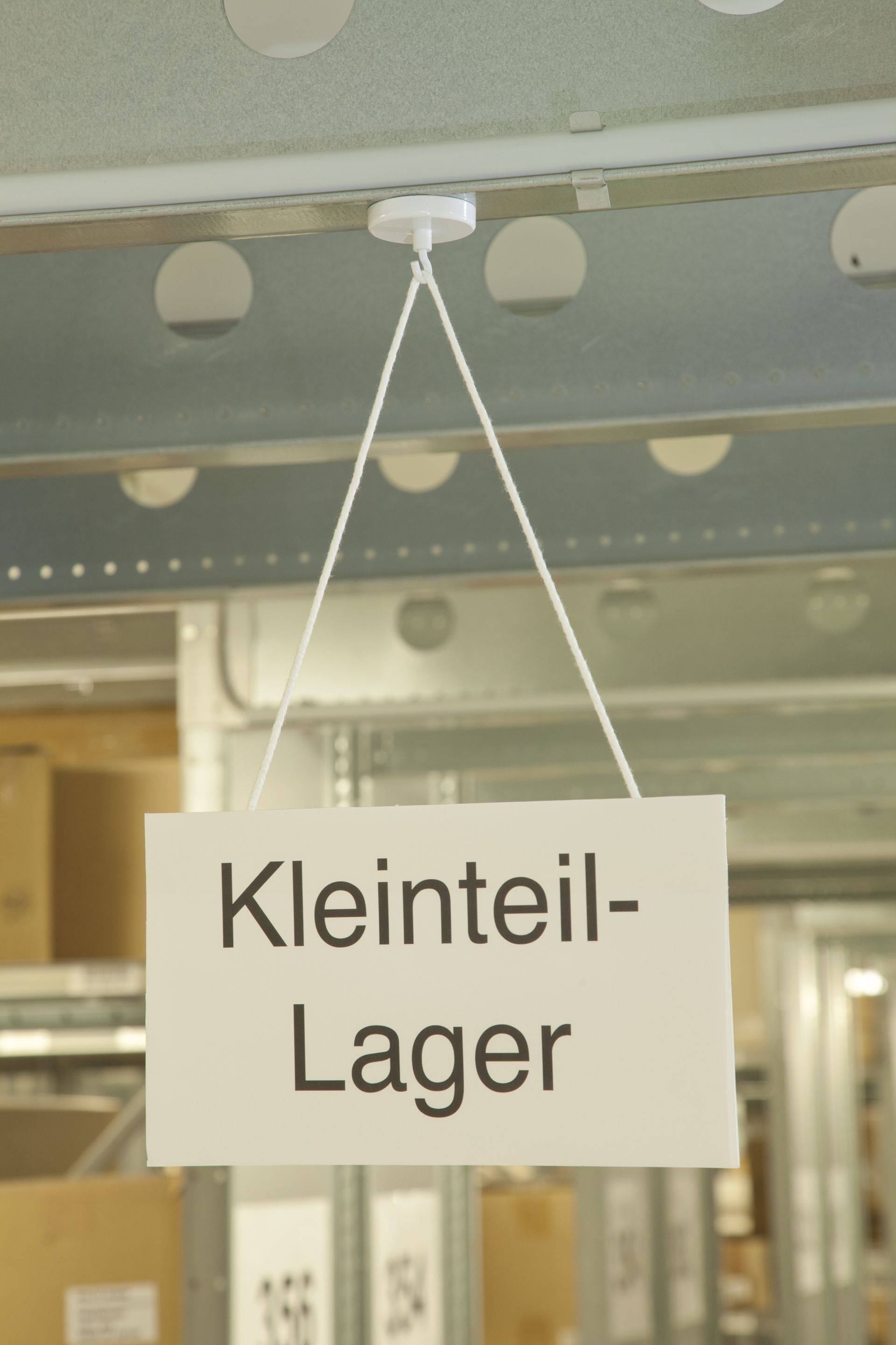 Ein weißes Schild mit dem deutschen Text 'Kleinteil-Lager' hängt an einem Metallträger in einer industriellen Umgebung und weist auf einen Kleinteile-Lagerbereich hin.