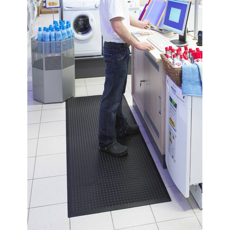 Eine Person in einem weißen Hemd und dunkler Hose steht an einem Tresen in einem Elektrogeschäft und bedient einen Computer. In der Nähe sind Waschmaschinen und Reinigungsprodukte ausgestellt.