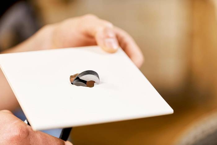 Eine Hand hält eine weiße rechteckige Karte mit einem kleinen, unregelmäßig geformten Loch in der Mitte, wodurch ein geschichtetes Objekt darunter sichtbar wird.