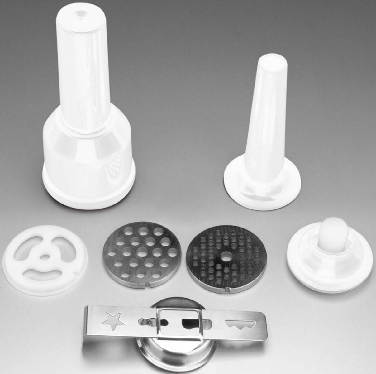 Eine Sammlung von Küchengeräte-Zubehörteilen, darunter Wurstfüller, Fleischwolf-Scheiben und ein Teil eines Nudelmakers vor einem grauen Hintergrund.