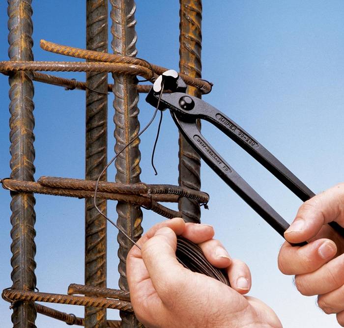 Hände, die mit einer Zange Draht um Metallbewehrungsstäbe wickeln, vor einem blauen Himmelshintergrund.